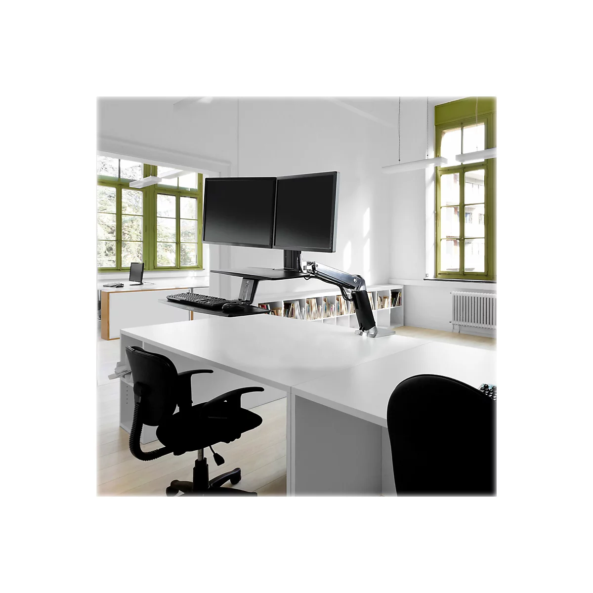 Büro mit zwei Monitoren auf einem verstellbaren Schreibtisch, Fenster mit grünen Rahmen.