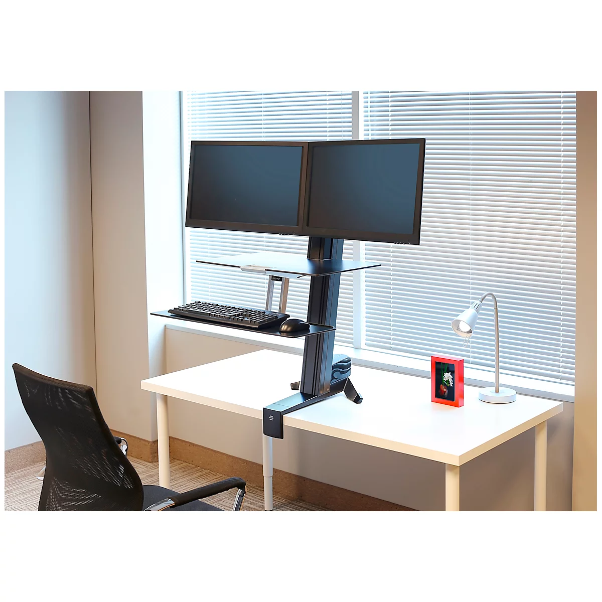 Office desk with two monitors, keyboard, and mouse. A lamp and a picture frame by the window.