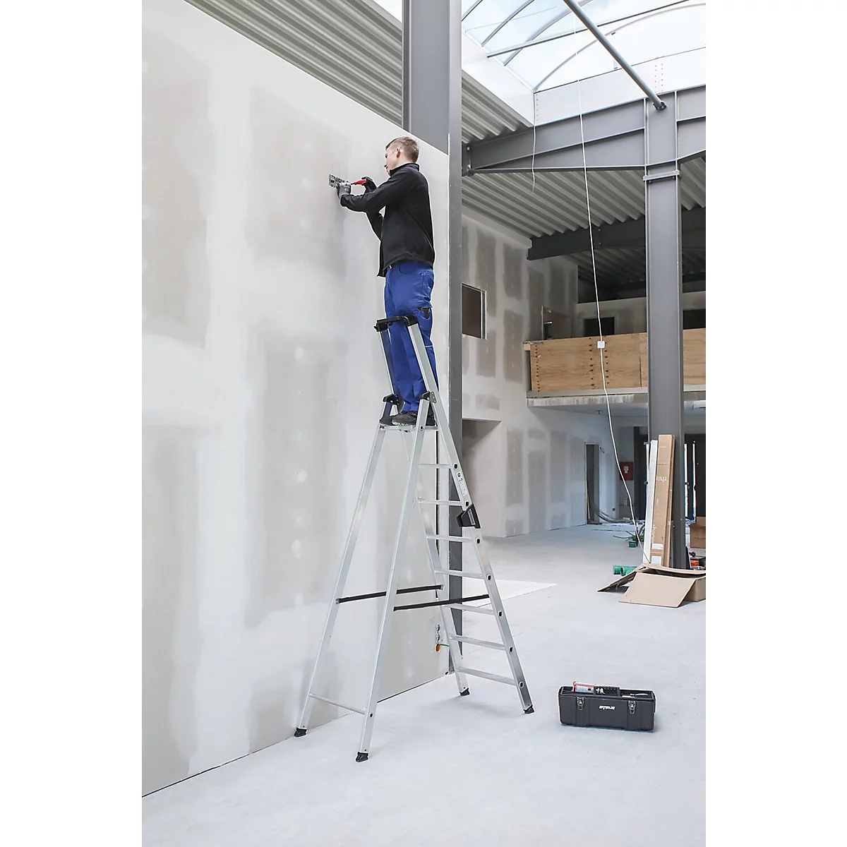 Un homme travaille sur un mur depuis une échelle. Boîte à outils au sol.