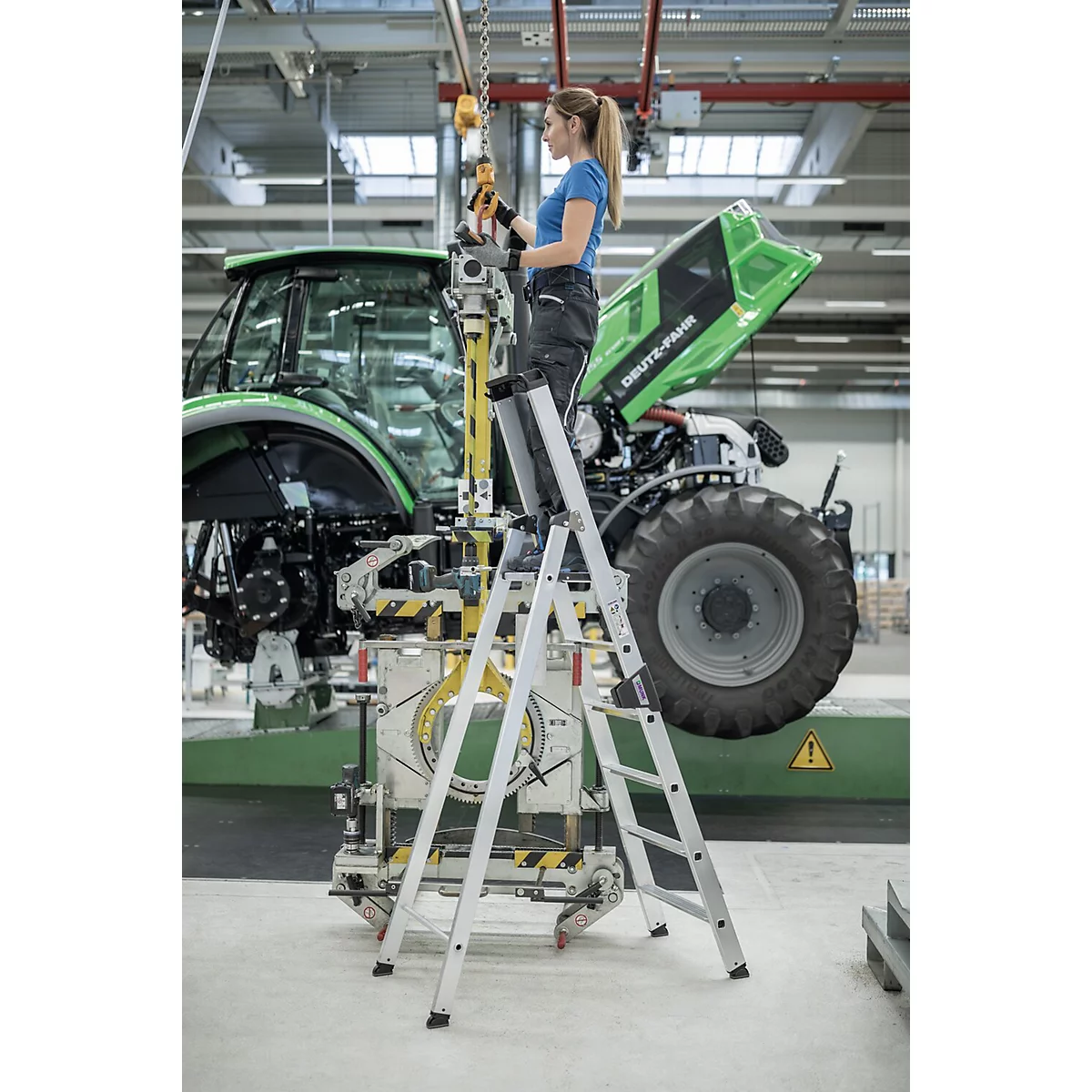 Une femme sur un escabeau travaille sur un tracteur dans une usine. Le tracteur porte l'inscription "DEUTZ-FAHR".