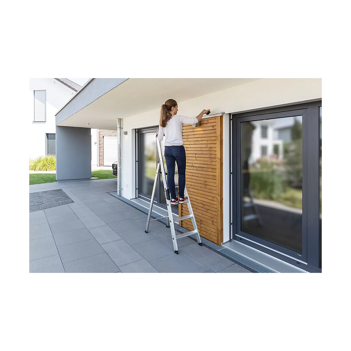 Une femme sur une échelle installe un écran d'intimité en bois. Le bâtiment a de grandes fenêtres et des carreaux gris.