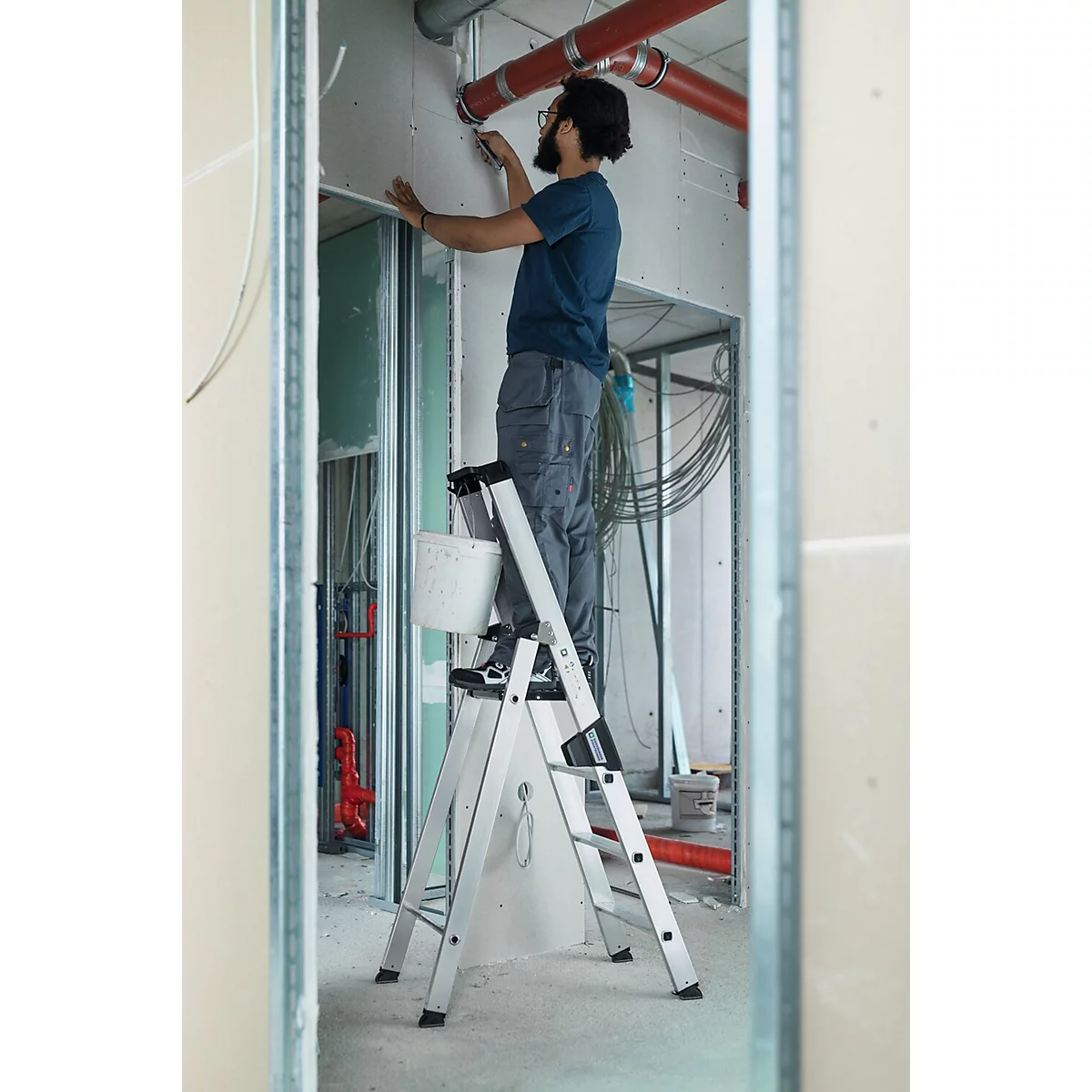 Homme sur une échelle lors de travaux de rénovation. Il peint un mur, avec des tuyaux et des câbles en arrière-plan.