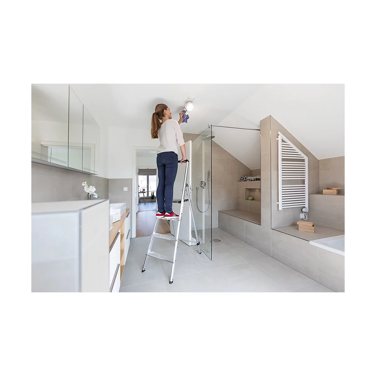 Femme sur une échelle nettoyant une lampe de plafond dans une salle de bain moderne.