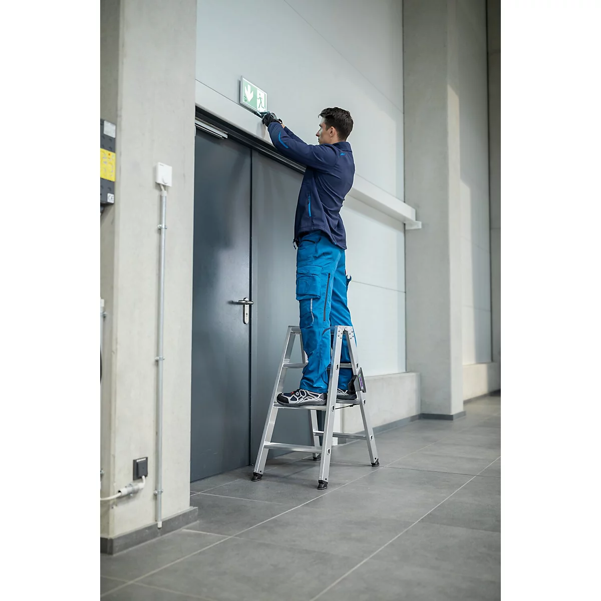 Un homme sur une échelle fixe un panneau de sortie de secours sur une porte.