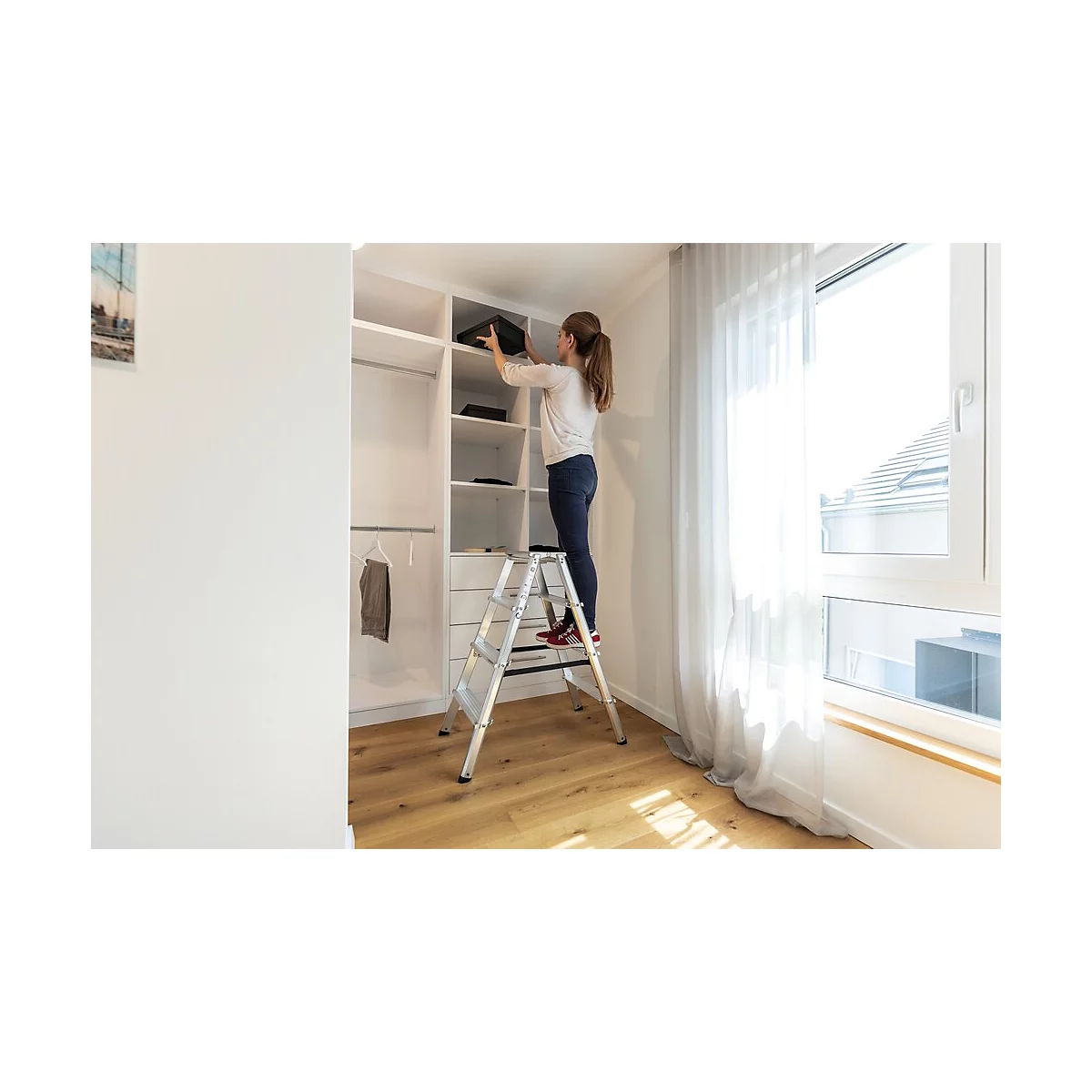 Une femme range dans un placard blanc, debout sur une échelle argentée. Pièce lumineuse avec fenêtre.