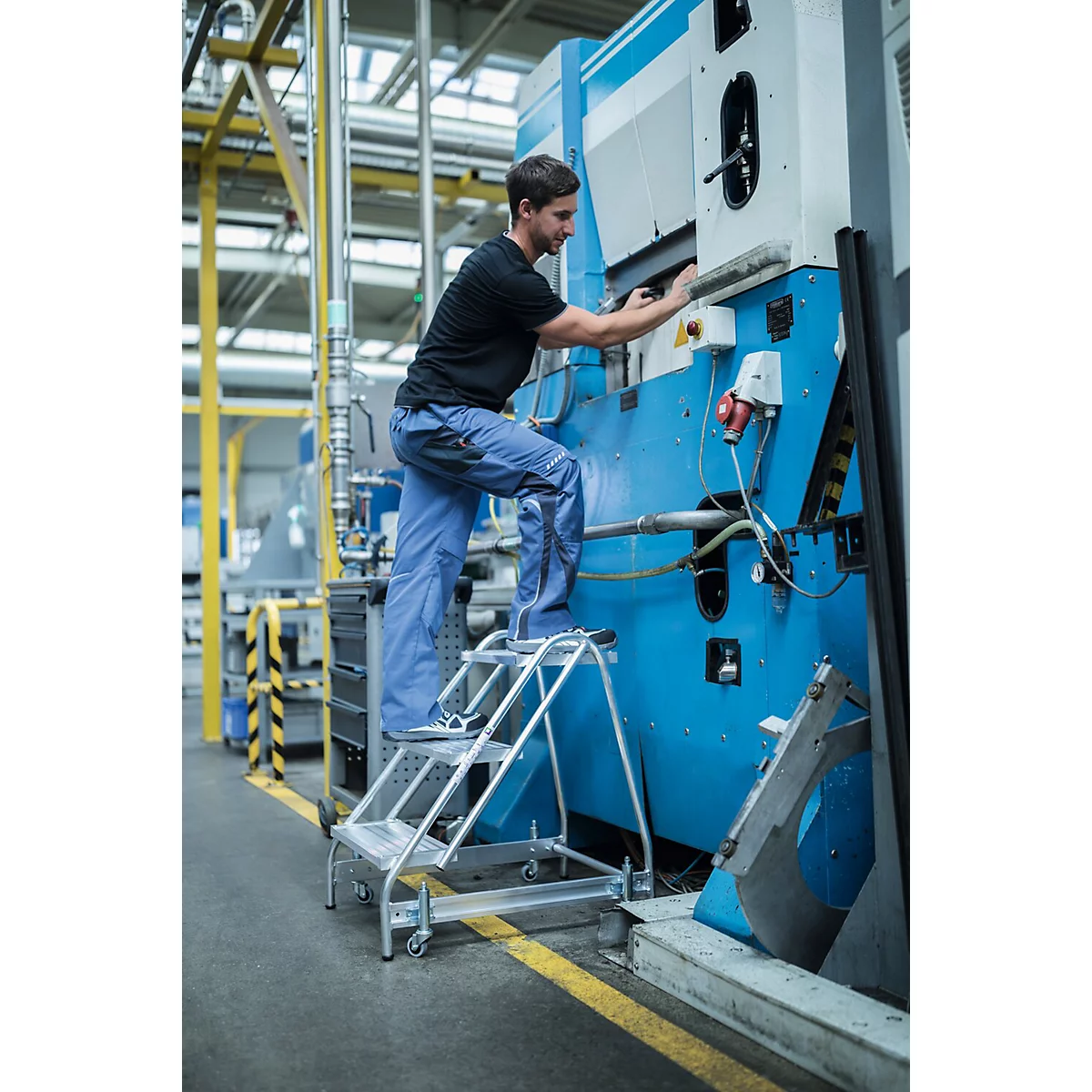 Un homme est debout sur un escabeau en train de travailler sur une grande machine bleue dans une usine.