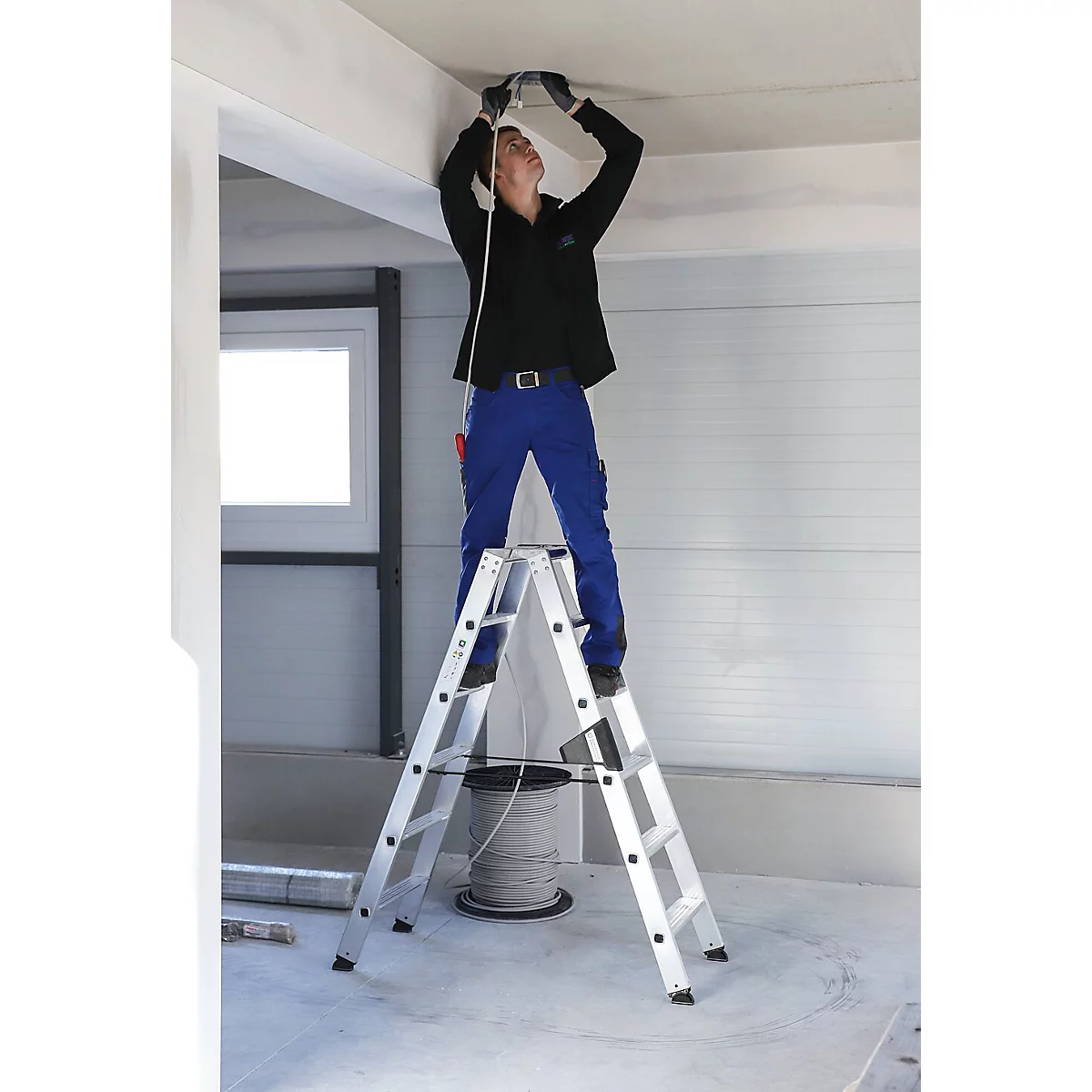 Un homme en tenue de travail bleue est debout sur une échelle, installant une lampe au plafond.