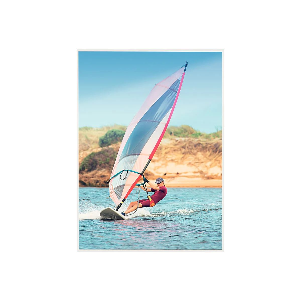Ein Windsurfer in roter Kleidung surft auf blauem Wasser vor einer sandigen Küste.