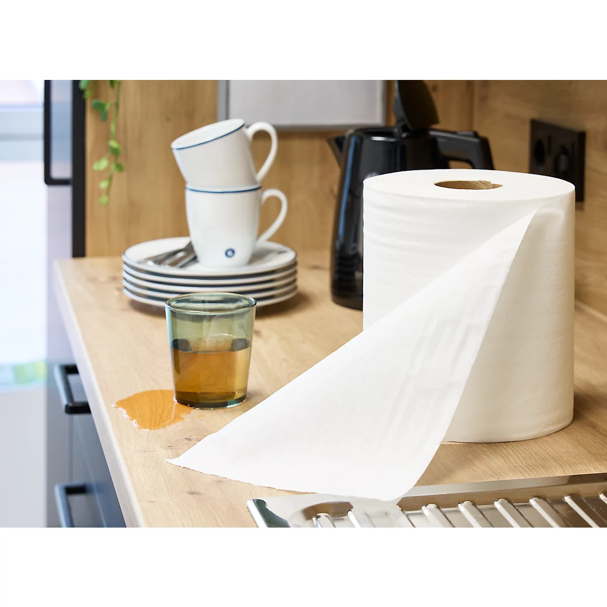 Une feuille d'essuie-tout est sur un comptoir de cuisine, à côté d'un verre, de tasses et d'assiettes. Il y a une tache de liquide sur le comptoir.