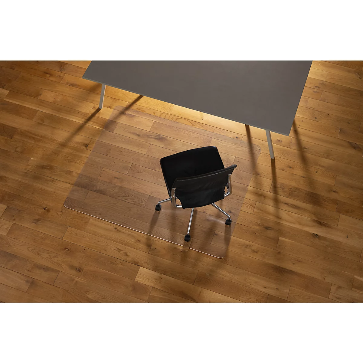 Silla de oficina sobre una alfombra protectora de suelo transparente debajo de una mesa gris sobre suelo de madera.