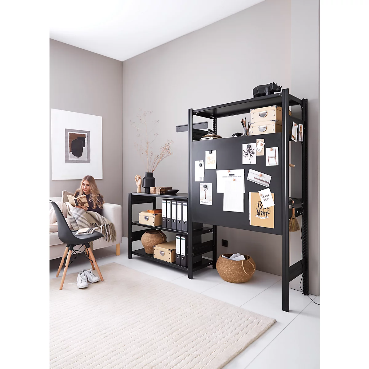 Une femme lit sur un canapé. Étagère noire avec notes et décor. Tapis clair, chaise.