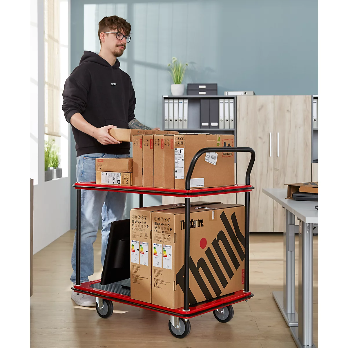 Een man met een bril laadt een rode transportwagen met pakketten. Er staan dozen en een monitor op de kar.