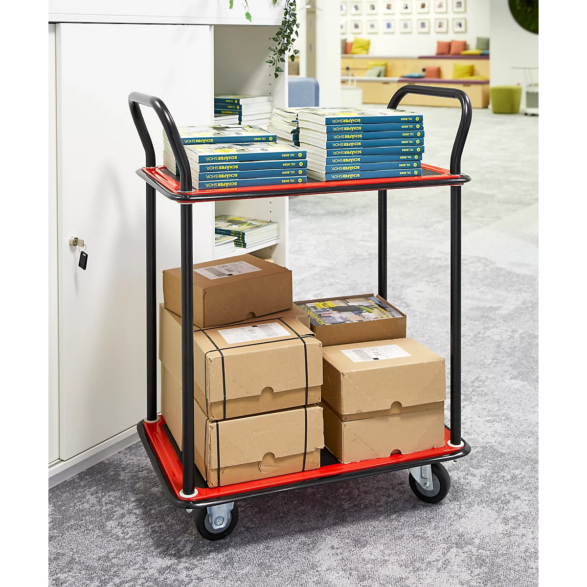 Een rode serveerwagen met boeken en pakketten op een kantoor. Op het bovenste niveau liggen boeken, dozen eronder. Zwart handvat.