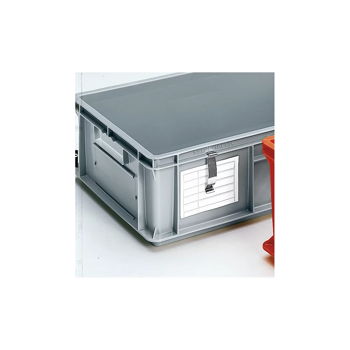 Grijze plastic box met deksel, adreslabel en metalen sluiting. Delen van een rode box zijn te zien op de achtergrond.