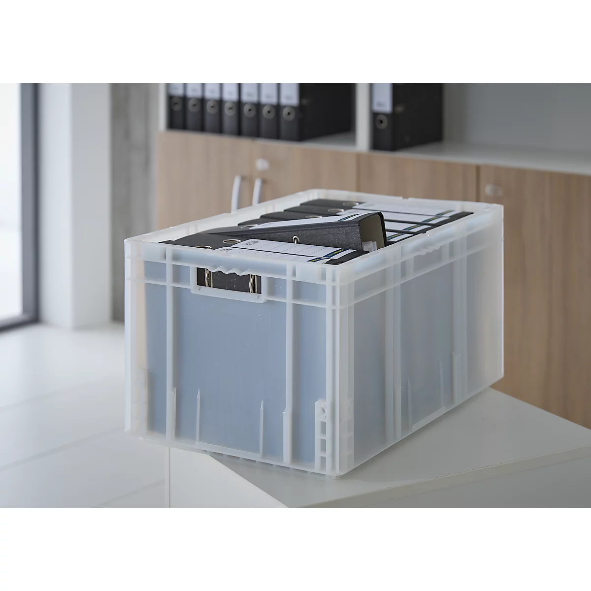 Boîte transparente avec des dossiers sur une étagère blanche, mobilier de bureau en arrière-plan.
