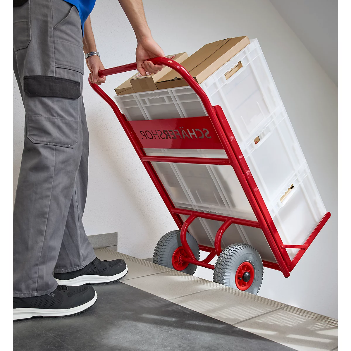 Person zieht eine rote Sackkarre mit Kisten und Kartons die Treppe hoch. Auf der Sackkarre steht "SCHWERER SHOP".