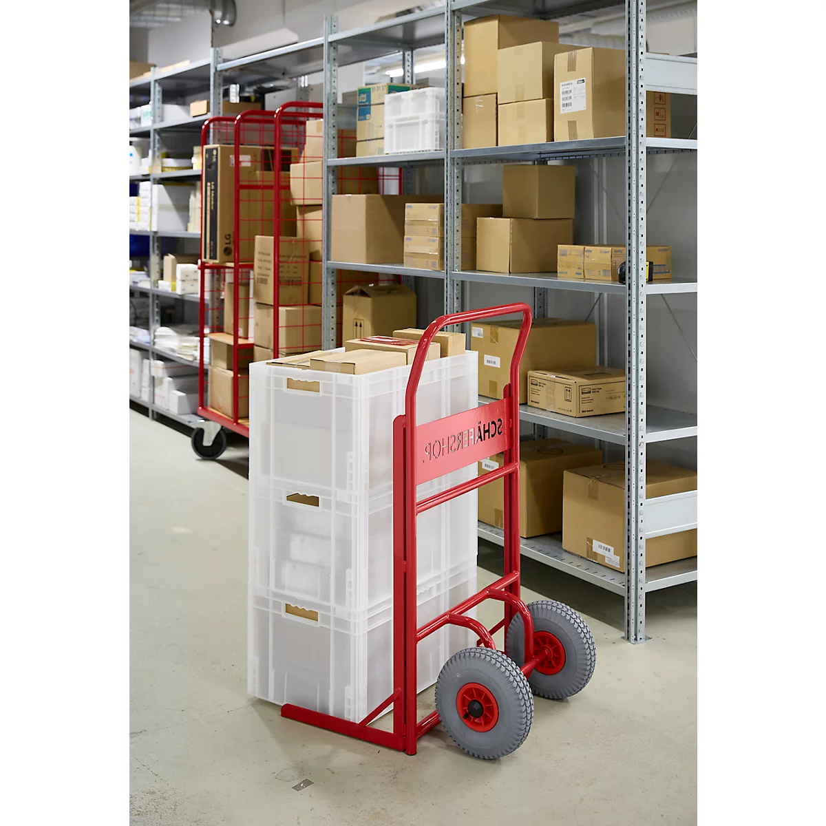Roter Transportwagen mit weißen Kisten in einem Lager; Regale mit Kartons im Hintergrund.