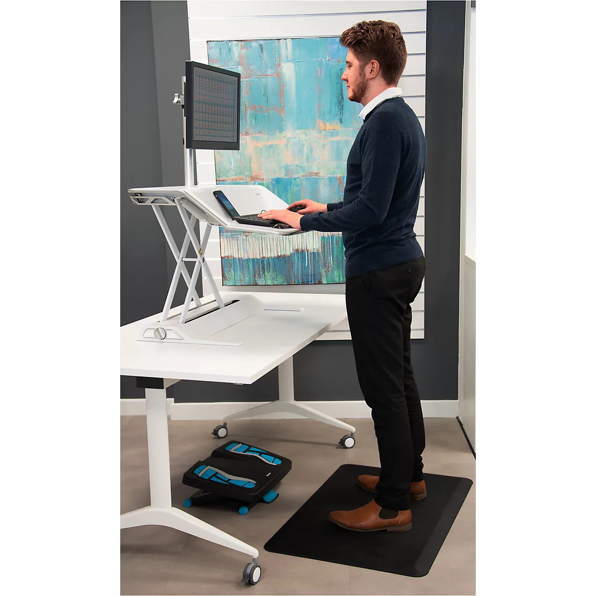 Man stands at a height-adjustable desk with laptop, monitor and footrest on a mat.