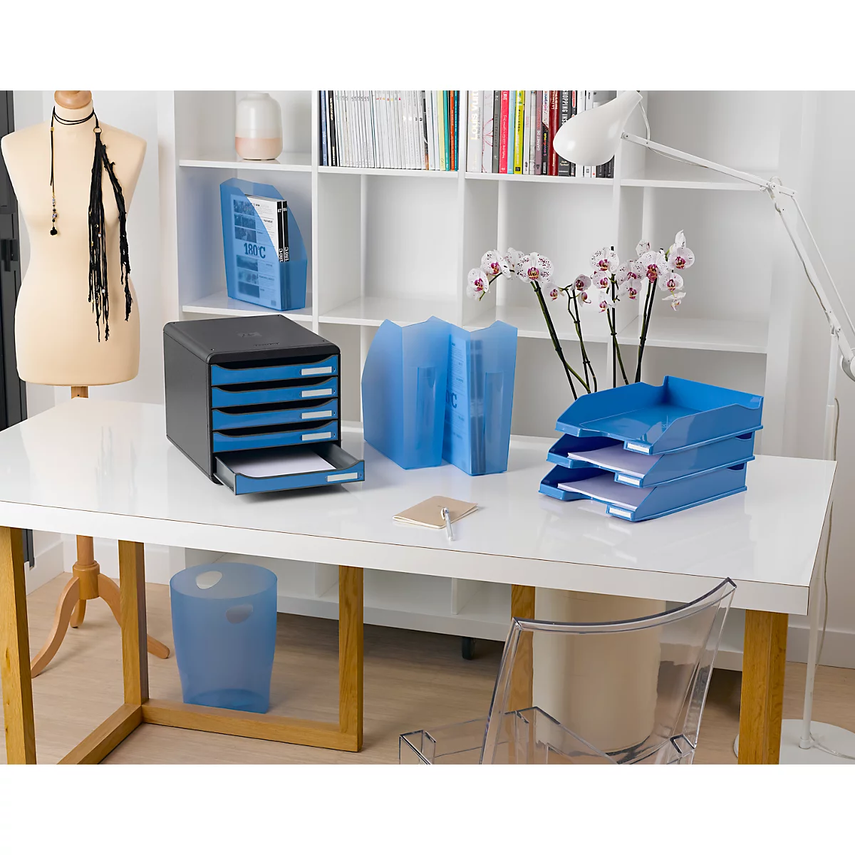 Bürotisch mit blauen Ablagen, Papierkorb, Ordnern und Dekoration. Heller Raum mit Schreibtisch und Bücherregal.