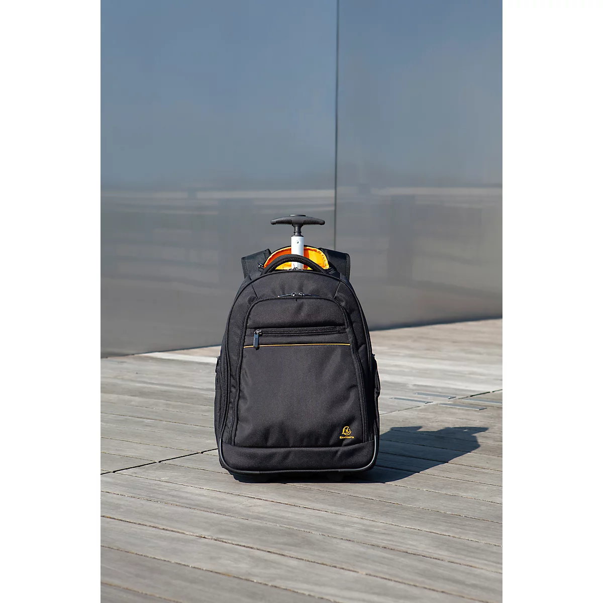 Schwarzer Trolley-Rucksack auf Holzuntergrund, vor einer grauen Wand.