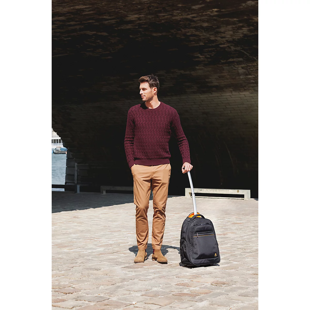 Ein Mann in weinrotem Pullover und beigefarbener Hose steht mit Rollkoffer. Er blickt zur Seite.
