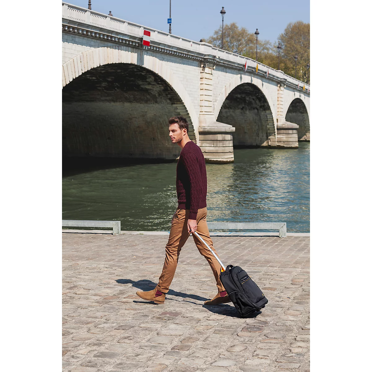 Ein Mann mit Koffer geht an einer Steinbrücke vorbei. Er trägt braune Hose, bordeauxroten Pullover und beige Schuhe.