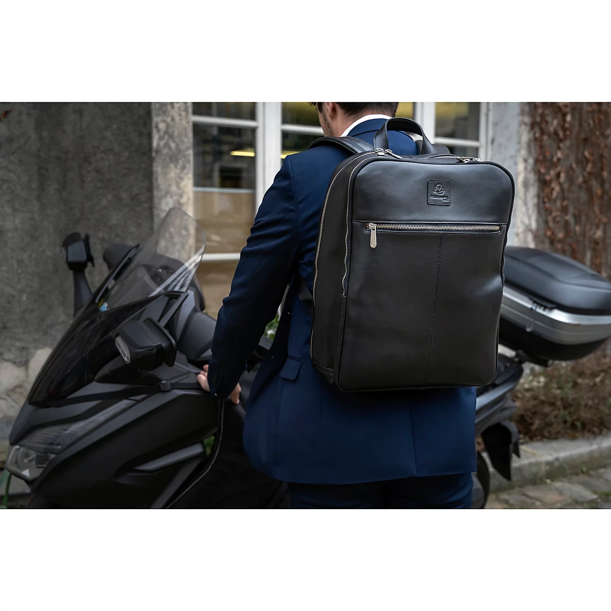 Ein Mann im Anzug mit einem schwarzen Lederrucksack steht neben einem Motorroller.