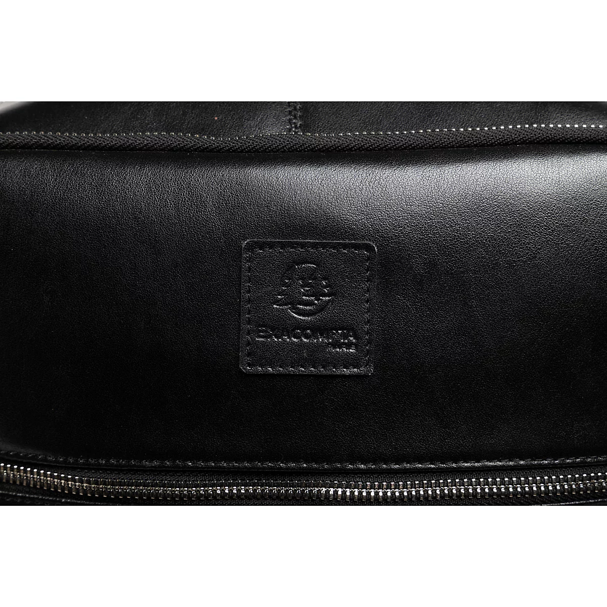 Detailaufnahme einer schwarzen Leder-Umhängetasche. Oben mit Ziernähten, unten mit Reißverschluss. Aufgenähtes Logo-Patch.