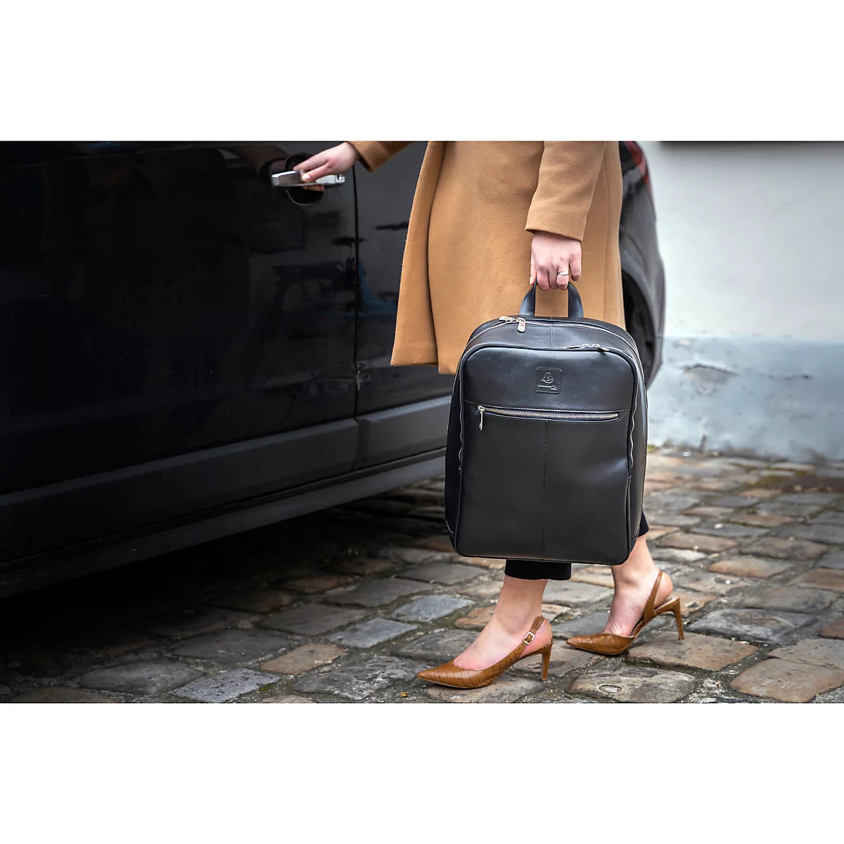 Eine Frau steigt mit einem schwarzen Rucksack in ein Auto. Sie trägt einen beigen Mantel und braune Schuhe.