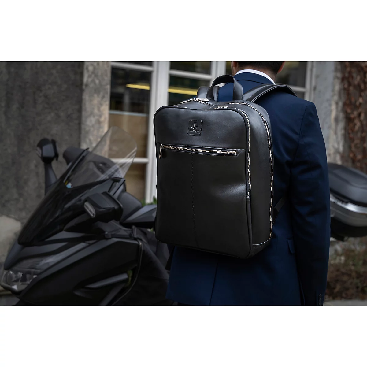 Ein Mann mit dunkelbraunem Lederrucksack geht neben einem schwarzen Motorrad.