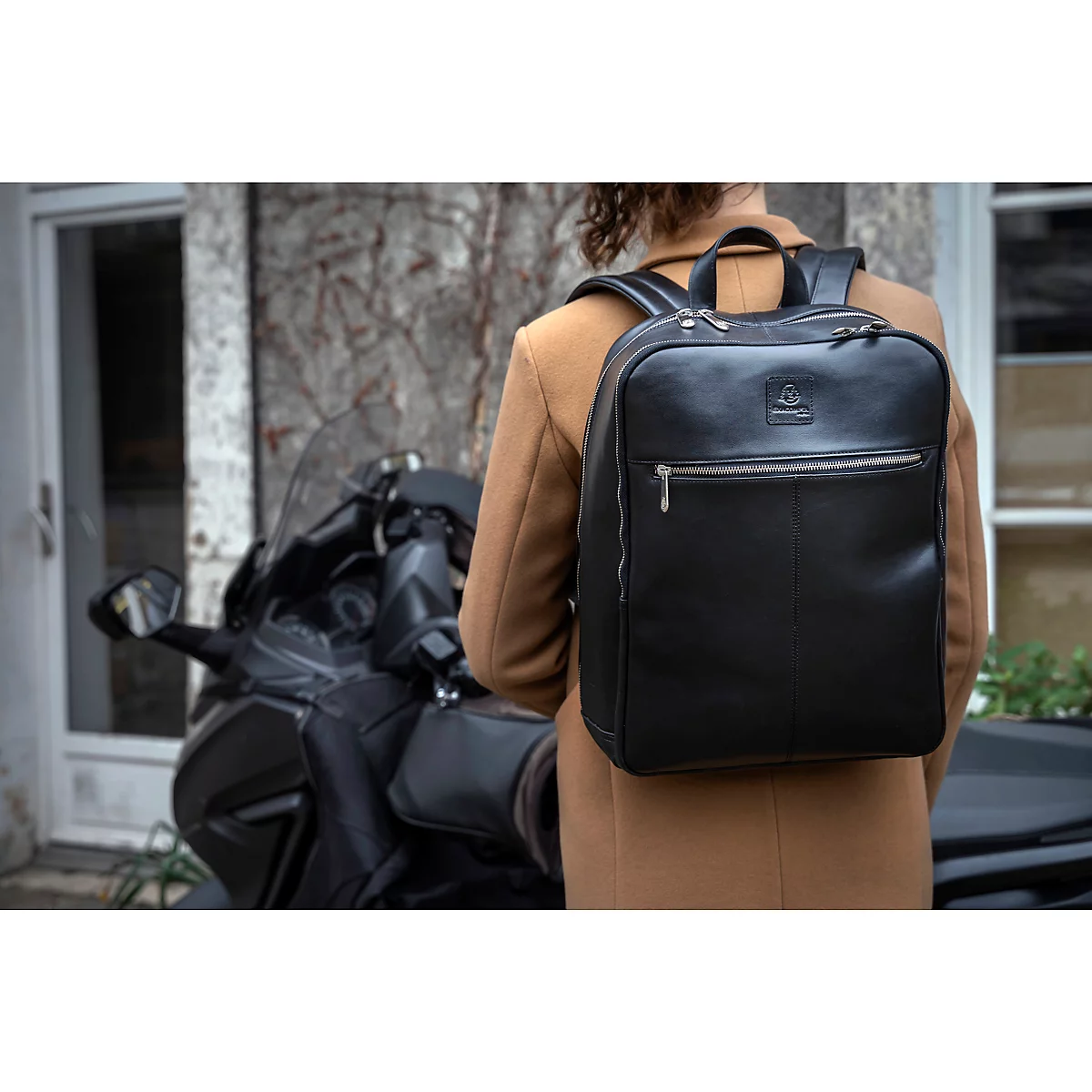 Une personne portant un sac à dos noir se tient à côté d'une moto. La personne porte un manteau beige.