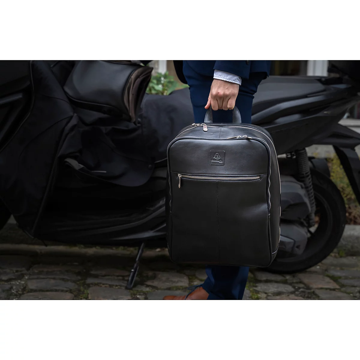 Une personne tient un sac à dos en cuir noir. En arrière-plan, un scooter, la personne porte une veste bleue.