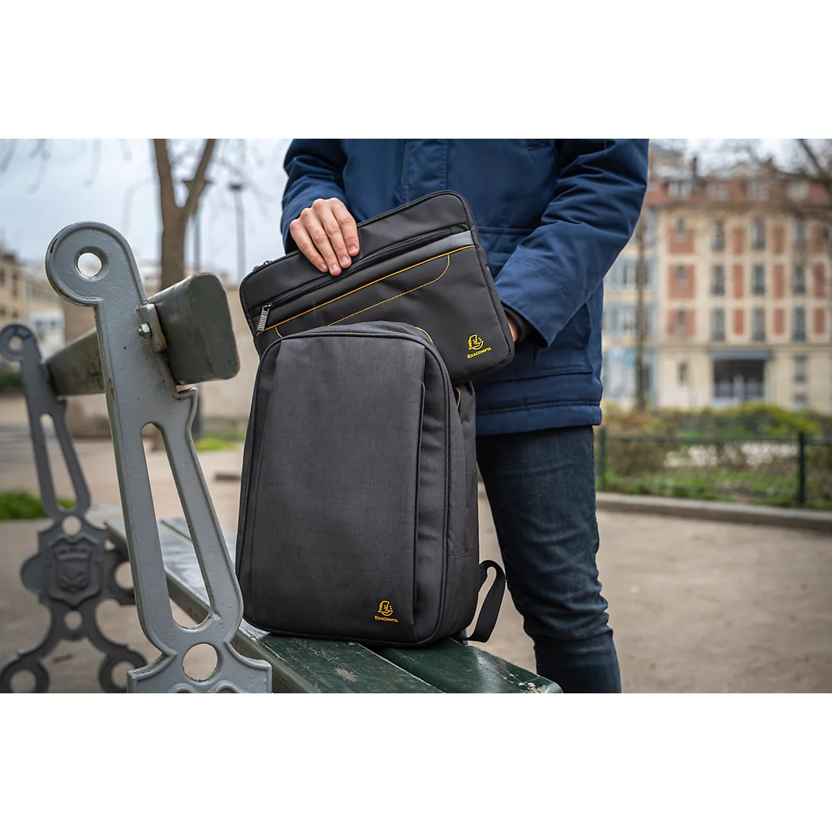 Mann hält Rucksack und Laptoptasche. Beide schwarz mit gelben Akzenten und Logo. Im Hintergrund Park mit Bank.