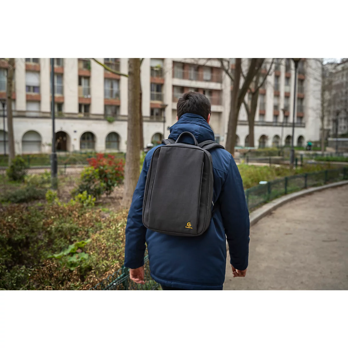Ein Mann mit schwarzem Rucksack geht einen Weg entlang, Gebäude im Hintergrund.