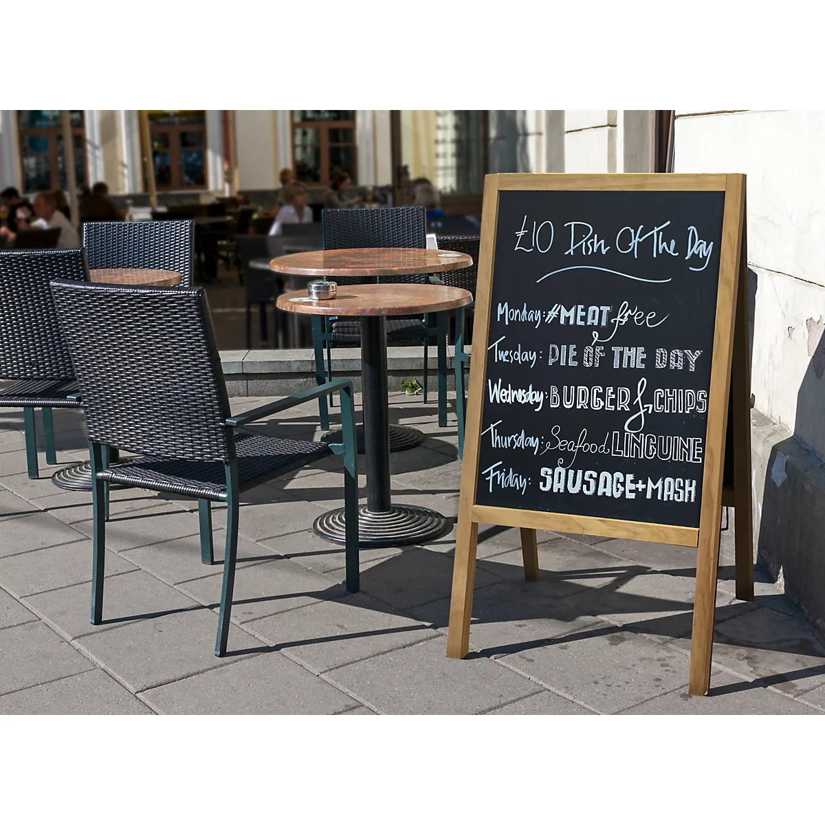 Ein Menü-Tafel vor einem Café, zeigt Tagesgerichte: Meat Free, Burger & Chips, usw. Draussen stehen Tische und Stühle.