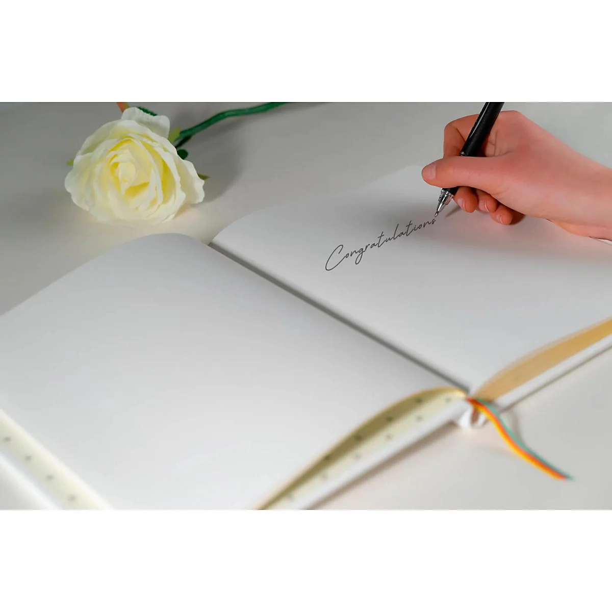 Gros plan sur un livre ouvert avec l'inscription manuscrite "Félicitations". Une main écrit sur une page avec un stylo, à côté se trouve une rose.