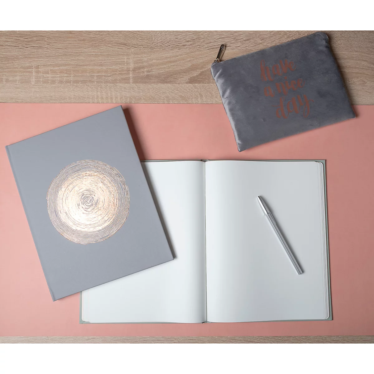 Cahier gris avec motif doré, à côté d'un cahier ouvert, un stylo argenté et une pochette grise.