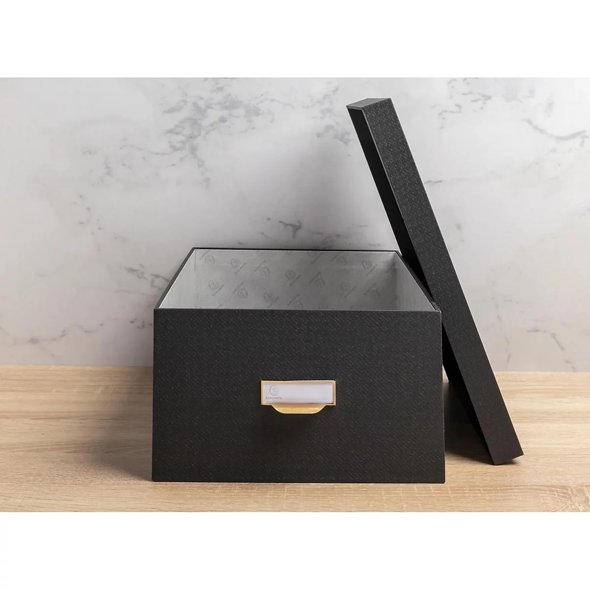 Boîte de rangement noire ouverte avec couvercle. Avec une étiquette blanche et une poignée dorée sur du bois. Fond marbré.