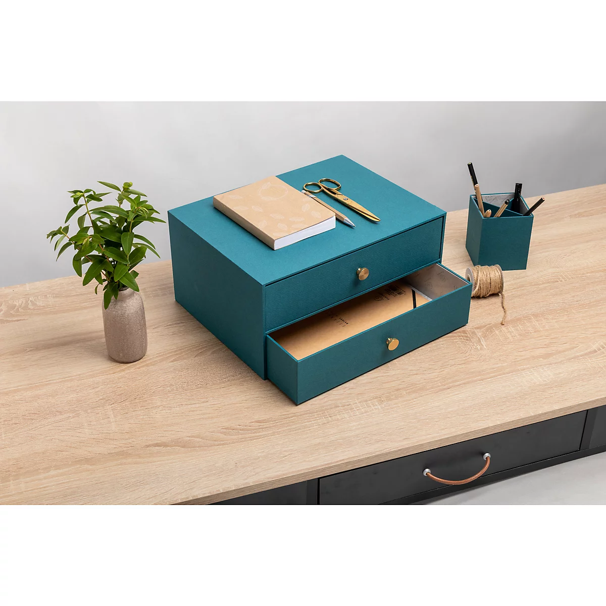 Tealfarbene Schubladenbox mit geöffneter Schublade, Schere, Notizblock, Stiftehalter und Pflanzen auf einem Holzschreibtisch.