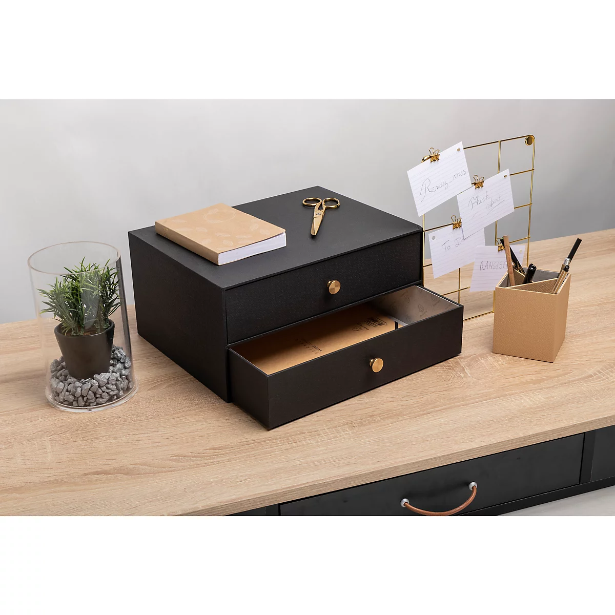 Schwarze Schreibtischablage mit goldfarbenen Griffen. Oben liegen Notizbuch und Schere. Auf hellem Holztisch, daneben Pflanze und Notizen.