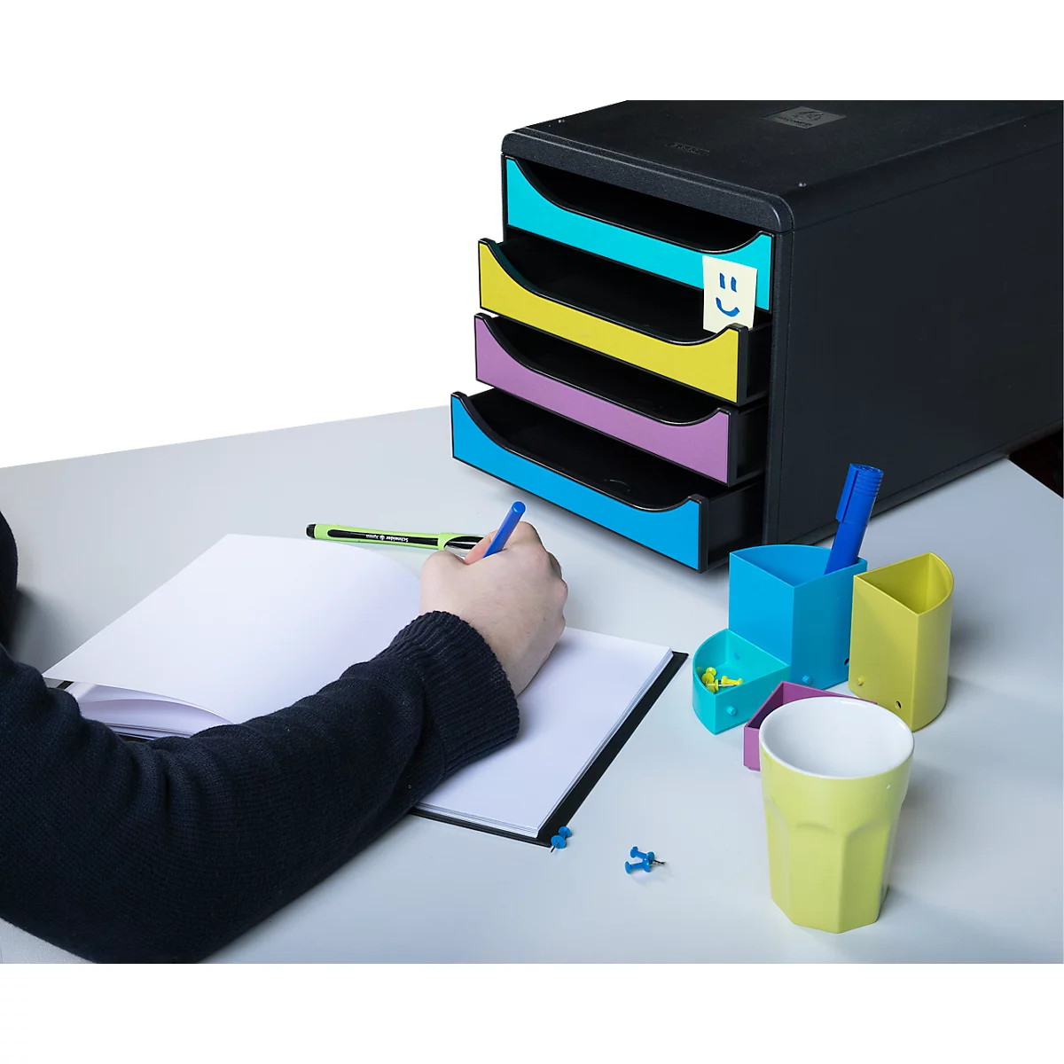 Personne écrivant sur un bloc-notes, à côté d'un système de classement noir avec des tiroirs colorés et des accessoires de bureau.