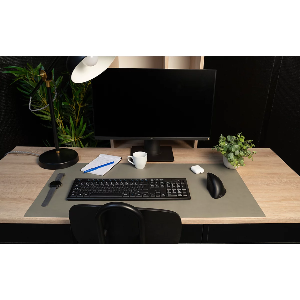 Schreibtisch mit Computer, Tastatur, Maus, Lampe und Pflanzen. Auf dem Schreibtisch liegt Papier und ein Becher.