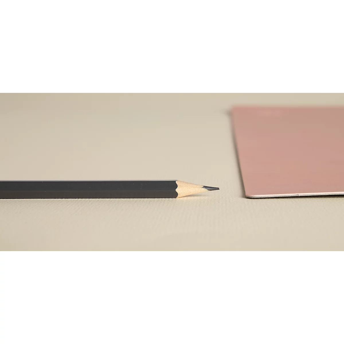Ein Bleistift liegt neben einem rosafarbenen Papier auf einem beigen Untergrund. Die Bleistiftspitze ist scharf.