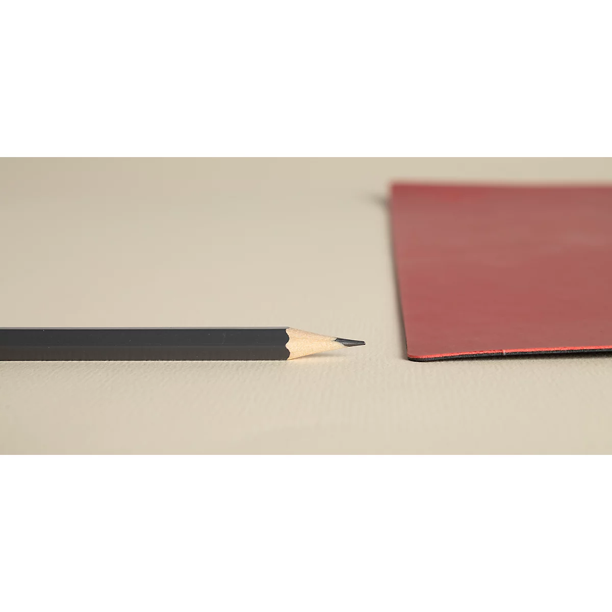 Ein schwarzer Bleistift liegt neben einem roten Papier auf einer hellen Oberfläche. Der Bleistift ist angespitzt.