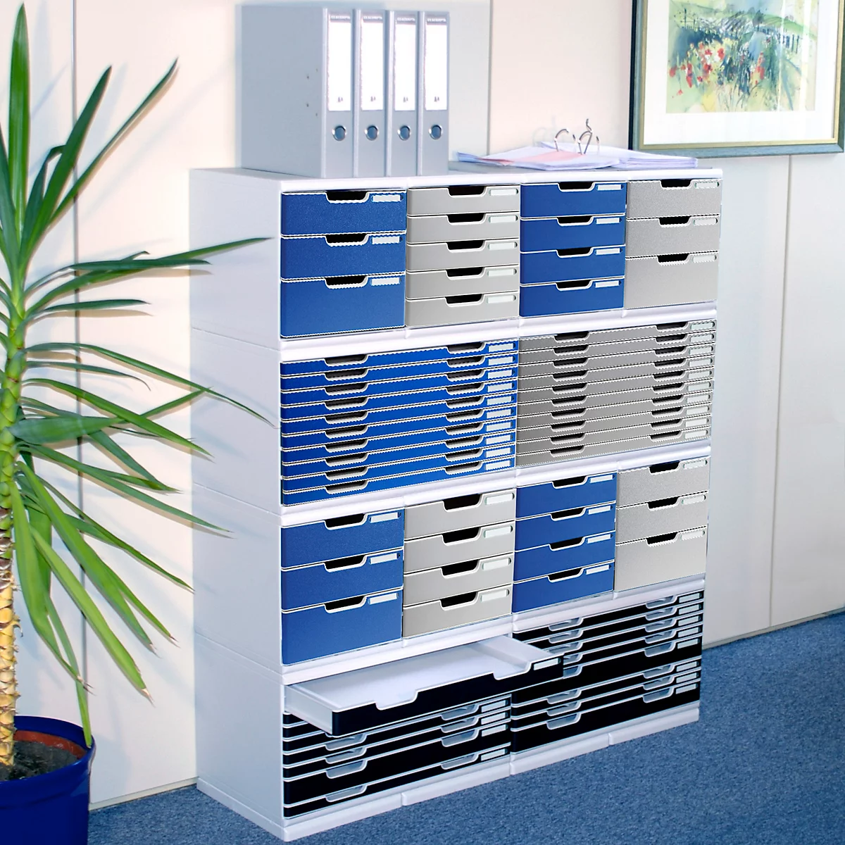 Weisses Büroregal mit Schubladen in Blau, Grau und Schwarz. Oben liegen Ordner, links eine Pflanze.