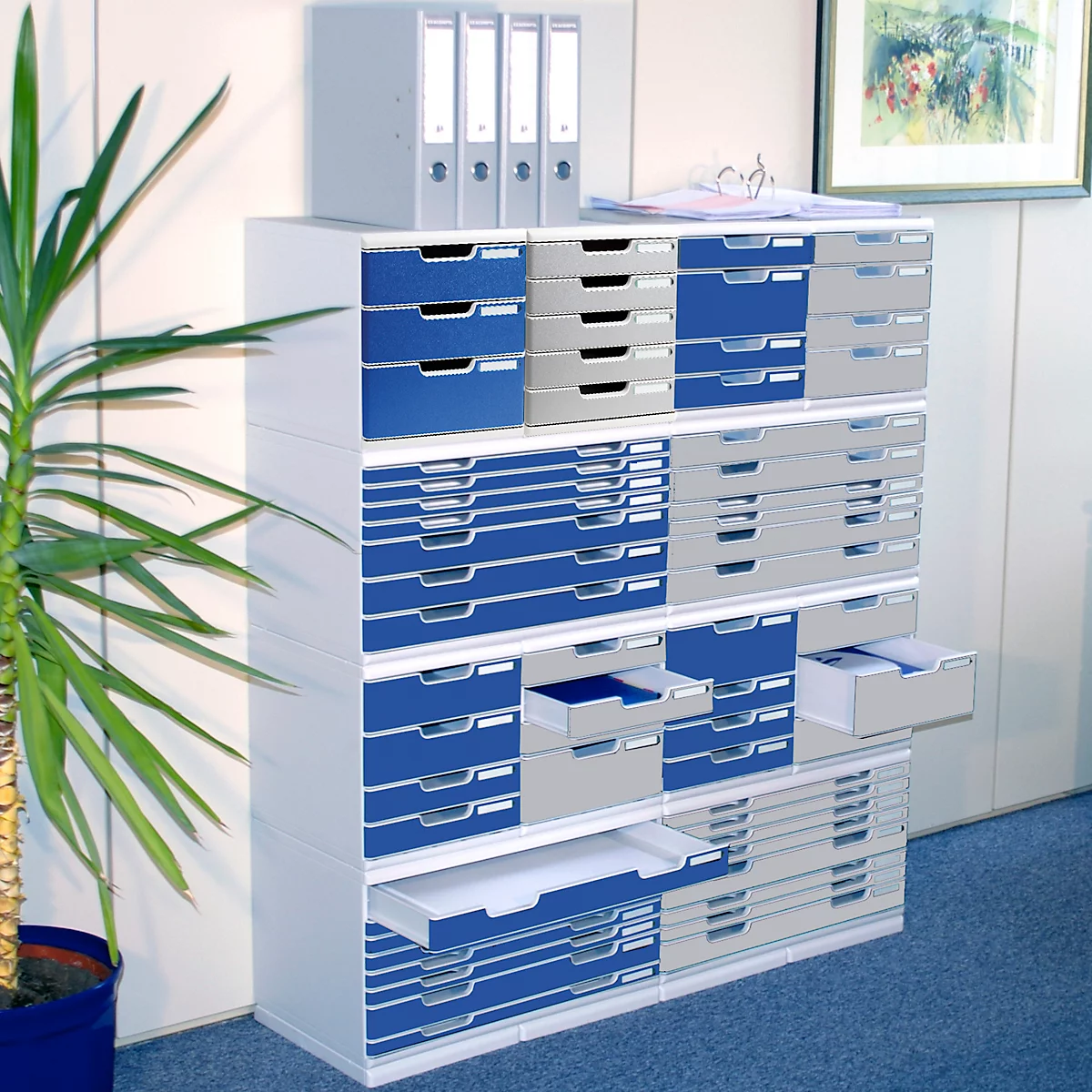 Ein Büroregal mit Schubladen, teilweise geöffnet. Blau-grau, Pflanzen und Ordner im Hintergrund.