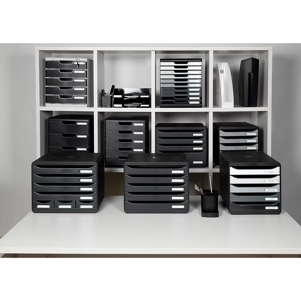 Schwarze Ablagesysteme mit weißen Etiketten auf einem weißen Schreibtisch und in einem Regal. Unterschiedliche Größen und Formen.