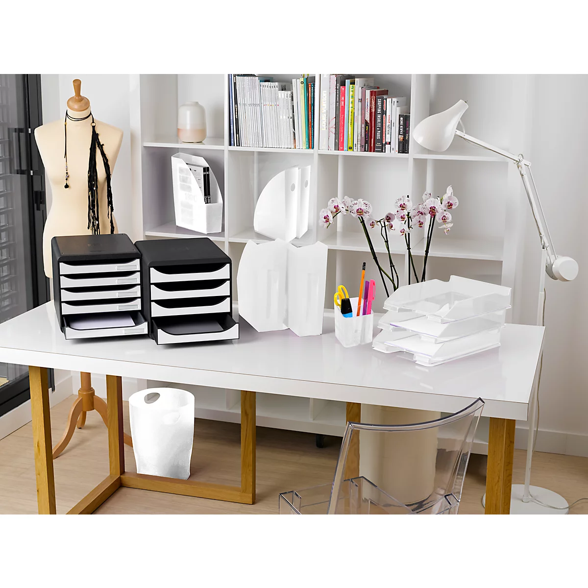 Schreibtisch mit schwarzen Ablagesystemen, weißer Tischlampe, Orchideen und Büromaterial.