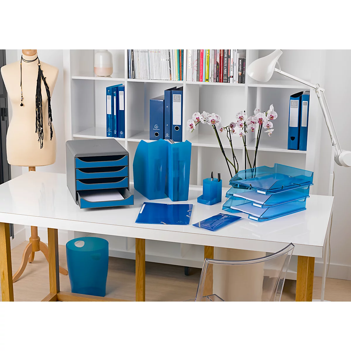 Büro-Set auf weissem Tisch. Blaue Ablagen, Ordner, Stifthalter, Papierkorb und Orchideen.