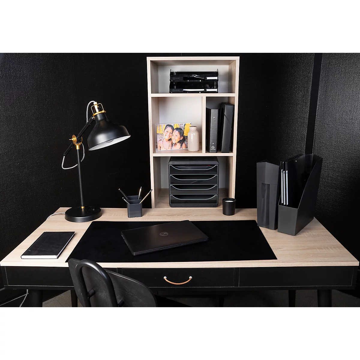 Schreibtisch mit Laptop, Lampe, Bücherregal, schwarzem Tischläufer und Stuhl. Dekoriert mit Zeitschriften, Bechern und Ordnern.